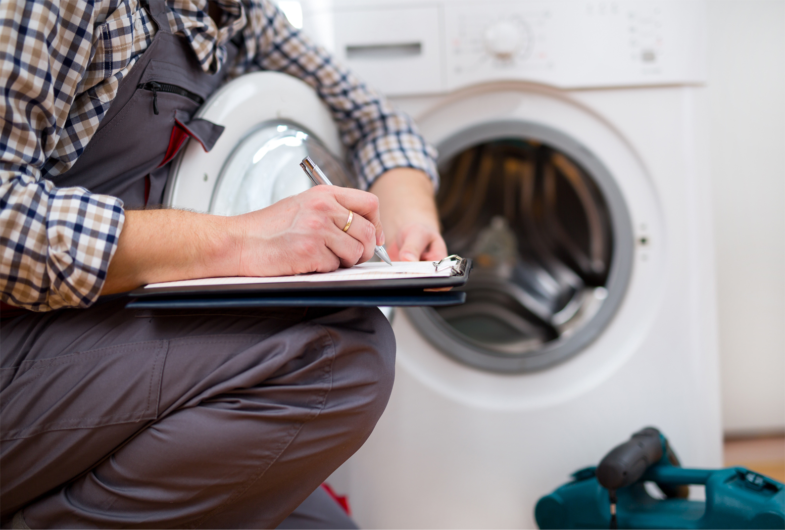 An appliance repair main crouching by a washing machine while writing on a clipboard